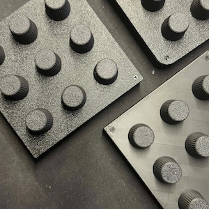 Op de afbeelding: Close-up van drie zwarte, getextureerde vierkante panelen, elk met negen gelijkmatig verdeelde, cilindrische knoppen. De knoppen hebben een geribbeld ontwerp. De panelen zijn met kleine schroeven in elke hoek bevestigd.