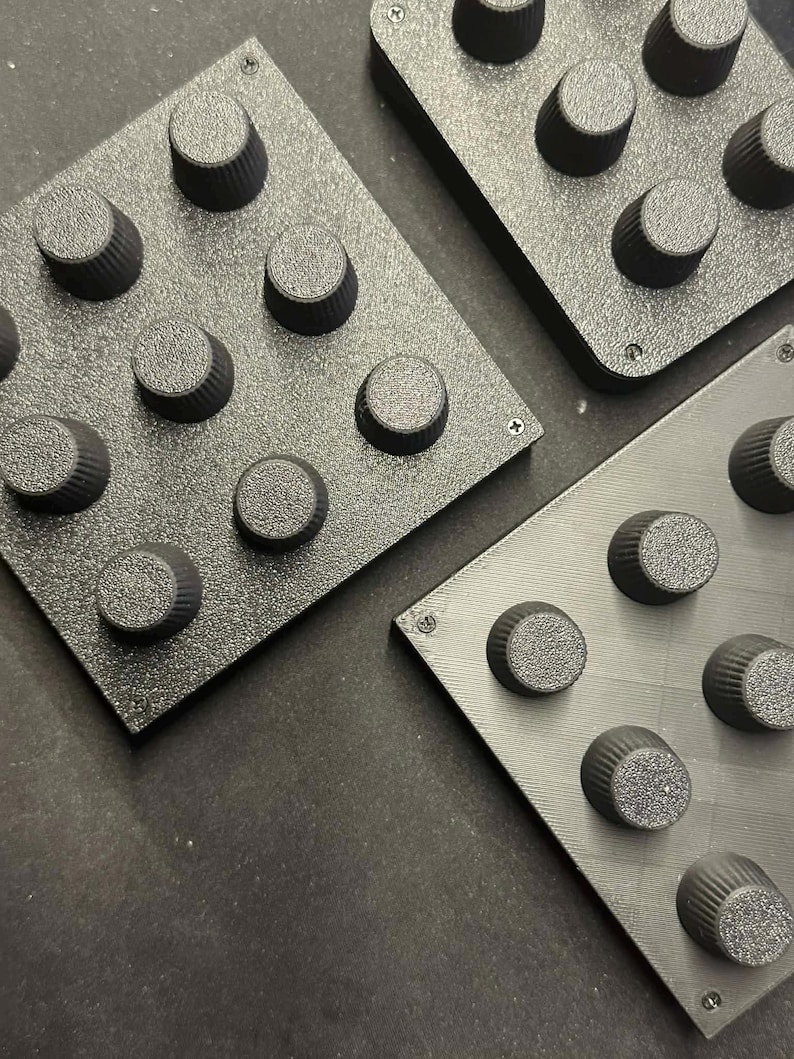 Op de afbeelding: Close-up van drie zwarte, getextureerde vierkante panelen, elk met negen gelijkmatig verdeelde, cilindrische knoppen. De knoppen hebben een geribbeld ontwerp. De panelen zijn met kleine schroeven in elke hoek bevestigd.