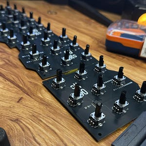 Op de afbeelding: Close-up van zwarte printplaten met talrijke kleine, zwarte knoppen en zilveren componenten. De platen zijn gerangschikt op een houten oppervlak, met een paar andere items zichtbaar op de achtergrond.