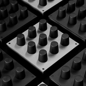 Puede incluir: Imagen en escala de grises que muestra múltiples paneles de control electrónicos cuadrados. Cada panel presenta una cuadrícula de perillas cilíndricas negras. Los paneles alternan entre negro y gris claro, creando un patrón repetitivo. La imagen es un primer plano, destacando la textura y el diseño.