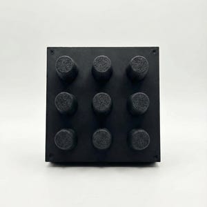 Op de afbeelding: Een zwart vierkant object met negen cilindrische knoppen gerangschikt in een 3x3 raster. De knoppen hebben een textuur. Het object staat tegen een effen witte achtergrond.