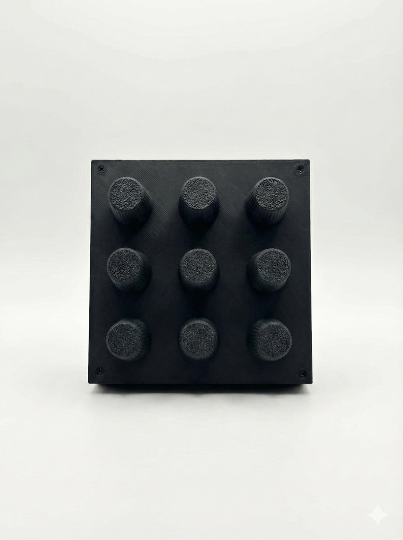 Op de afbeelding: Een zwart vierkant object met negen cilindrische knoppen gerangschikt in een 3x3 raster. De knoppen hebben een textuur. Het object staat tegen een effen witte achtergrond.