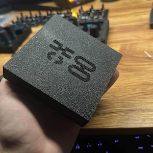 Op de afbeelding: Een zwarte, getextureerde vierkante doos met de tekst "CH 00" in reli&euml;f op de bovenkant. De doos wordt in de hand van een persoon gehouden, met een toetsenbord en computermonitor op de achtergrond.