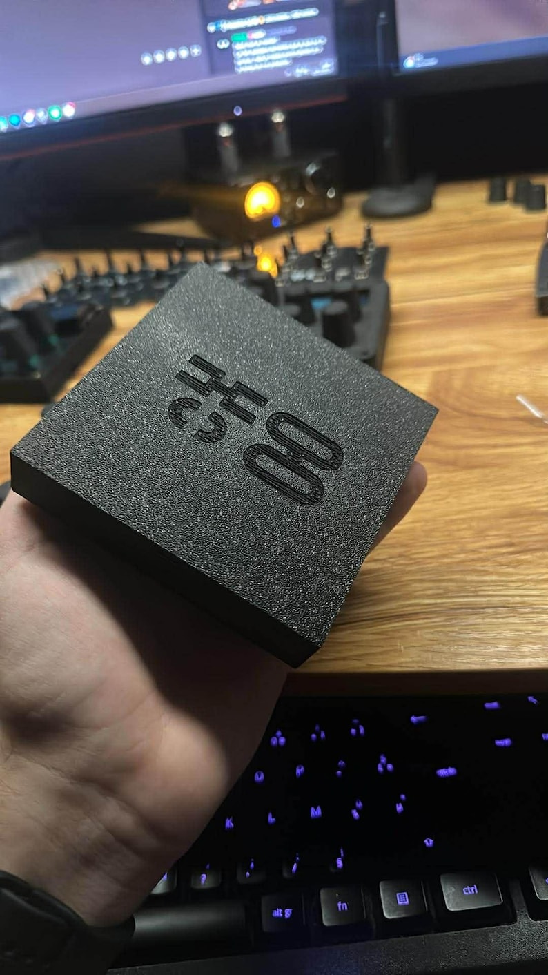 Op de afbeelding: Een zwarte, getextureerde vierkante doos met de tekst "CH 00" in reli&euml;f op de bovenkant. De doos wordt in de hand van een persoon gehouden, met een toetsenbord en computermonitor op de achtergrond.