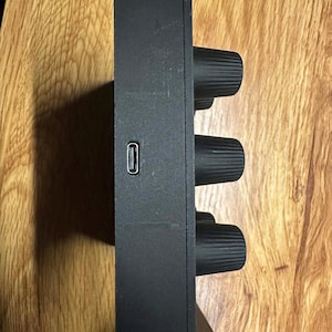 Op de afbeelding: Een zwart elektronisch apparaat met drie grote, getextureerde knoppen en een USB-poort. Het rechthoekige apparaat heeft een matte afwerking. De knoppen zijn gelijkmatig verdeeld aan de zijkant.