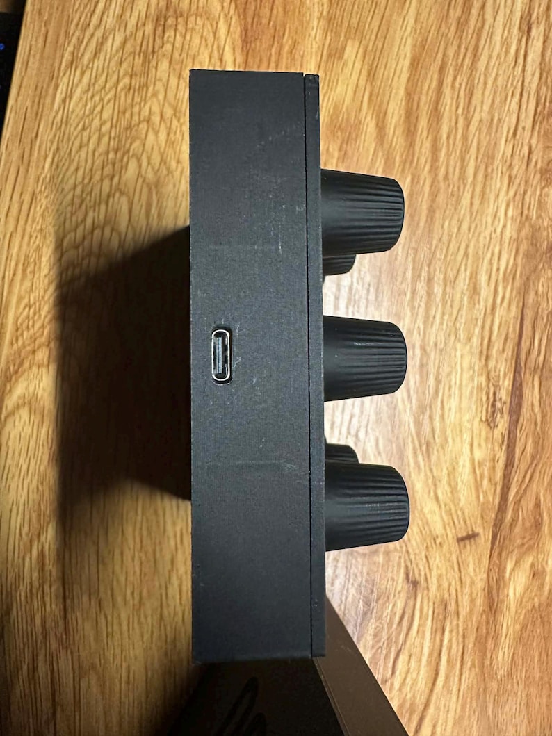 Op de afbeelding: Een zwart elektronisch apparaat met drie grote, getextureerde knoppen en een USB-poort. Het rechthoekige apparaat heeft een matte afwerking. De knoppen zijn gelijkmatig verdeeld aan de zijkant.