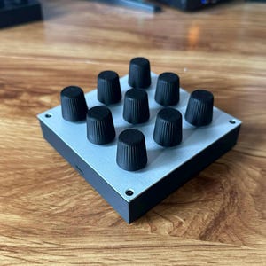 May include: A small, square electronic device with a light gray top and black sides. Nine black knobs are arranged in a 3x3 grid on the top surface. The device sits on a wooden surface.