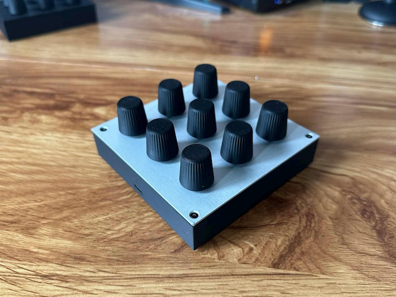 May include: A small, square electronic device with a light gray top and black sides. Nine black knobs are arranged in a 3x3 grid on the top surface. The device sits on a wooden surface.