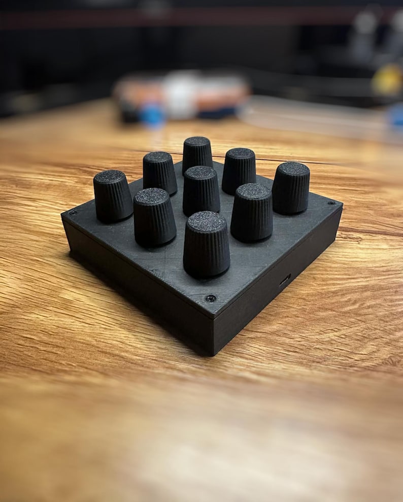 May include: A black electronic control panel with nine cylindrical knobs arranged in a 3x3 grid. The knobs have a textured surface. The panel is rectangular and sits on a wooden surface.