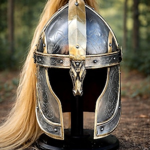 May include: An ornate silver and gold helmet with detailed patterns and a long, blonde hairpiece. The helmet has a decorative animal head at the front and is displayed on a black stand, set against a blurred forest background.