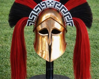 May include: A gold-colored Spartan helmet with a red and black plume. The helmet is displayed on a black stand against a green background. The plume features alternating red and black sections.