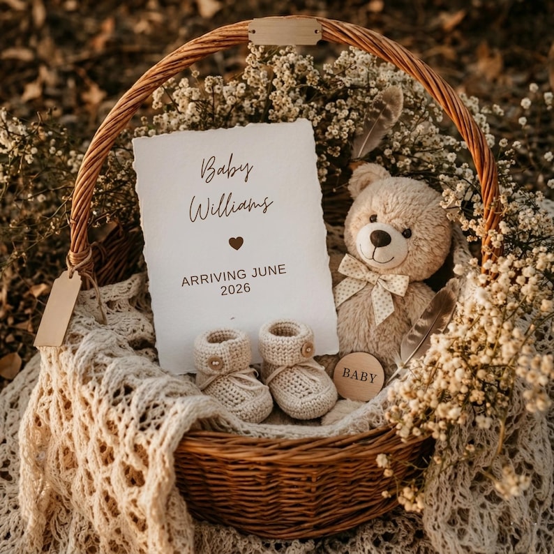 Rustic baby announcement basket featuring a plush teddy bear, cream knit booties, baby's breath flowers, and a card reading "Baby Williams Arriving June 2026" on a beige macrame blanket.
