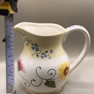 May include: White ceramic pitcher with floral designs, including sunflowers and pink flowers. The pitcher has a yellow band with blue floral accents. The pitcher is approximately 7 inches tall.