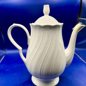 May include: A white porcelain teapot featuring a subtle, swirling embossed pattern across its body. The teapot includes a matching lid with a finial, a curved handle, and a spout with a small protective plastic cover attached to the tip.
