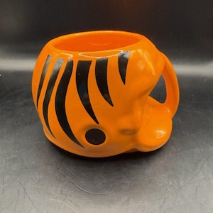 May include: Orange ceramic mug with black stripes and a handle. The mug has a rounded shape and a single black dot on the side. The mug is likely for beverages.