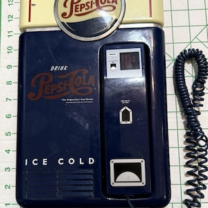 Puede incluir: Máquina expendedora de Pepsi-Cola vintage con la parte superior de color crema y el cuerpo azul oscuro. La máquina presenta el logotipo y el texto de Pepsi-Cola, "ICE COLD", y una ranura para monedas. Un cable azul en espiral está adjunto.