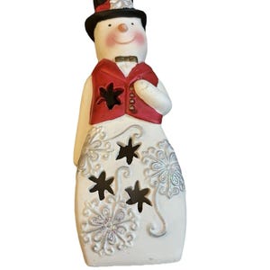 May include: A ceramic snowman figurine wearing a black top hat, red vest, and bow tie. The snowman has a carrot nose, rosy cheeks, and a smiling mouth. The body features snowflake and star cutouts.