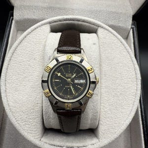 May include: A silver-toned CITIZEN 2000 quartz watch with a black dial, gold accents, and a brown leather strap. The watch is displayed in a gray, velvet-lined box. The watch face displays the day and date.