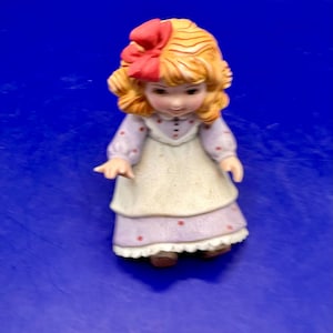 May include: A porcelain figurine of a young child with curly blonde hair styled with a large red bow. The figure wears a light purple long-sleeved dress featuring a small red polka dot pattern, paired with a cream-colored apron and a white ruffled hem.
