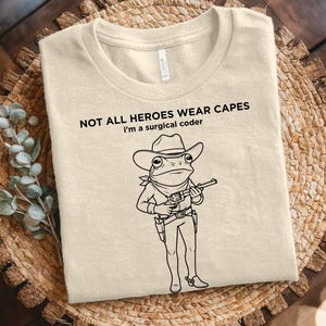 May include: A natural-colored t-shirt with the text "NOT ALL HEROES WEAR CAPES I'm a surgical coder." A cartoon frog wearing a cowboy hat and holding a gun is printed on the front. The shirt is displayed on a woven basket.
