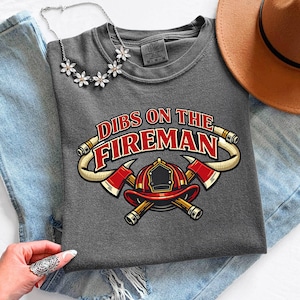 May include: A gray t-shirt with the text "Dibs on the Fireman" and a graphic of a firefighter's helmet and crossed axes. The shirt is laid on top of blue jeans, with a brown hat and a floral necklace.