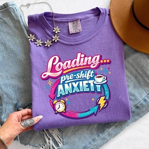 May include: A violet t-shirt with the text "Loading... pre-shift ANXIETY" in a colorful design. The shirt is paired with blue jeans, a brown hat, and a floral necklace. The shirt has a round neck.