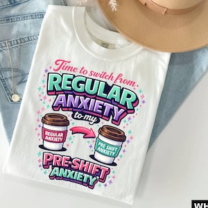 May include: White t-shirt with the text "Time to switch from REGULAR ANXIETY to my PRE-SHIFT ANXIETY." The design includes coffee cup graphics and colorful accents. The shirt is paired with blue jeans and a tan hat.