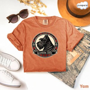 May include: A burnt orange t-shirt featuring a circular graphic of a black Scottish Terrier dog with the text "scottie life" on a banner. The graphic is framed by a red and black plaid pattern. The shirt is styled with sunglasses, a hat, and jeans.