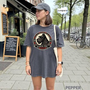 May include: Dark grey t-shirt featuring a circular design with a black Scottie dog illustration, red plaid accents, and the text "scottie life". The shirt is worn with a grey cap and a white tote bag.