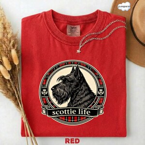 May include: A red t-shirt featuring a black and white illustration of a Scottish Terrier dog within a circular design. The design includes a plaid pattern and the words "scottie life". The shirt is made of Comfort Colors material.