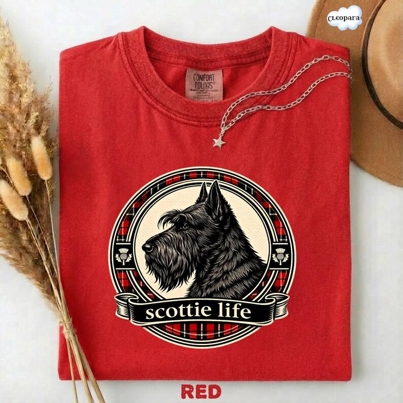 May include: A red t-shirt featuring a black and white illustration of a Scottish Terrier dog within a circular design. The design includes a plaid pattern and the words "scottie life". The shirt is made of Comfort Colors material.