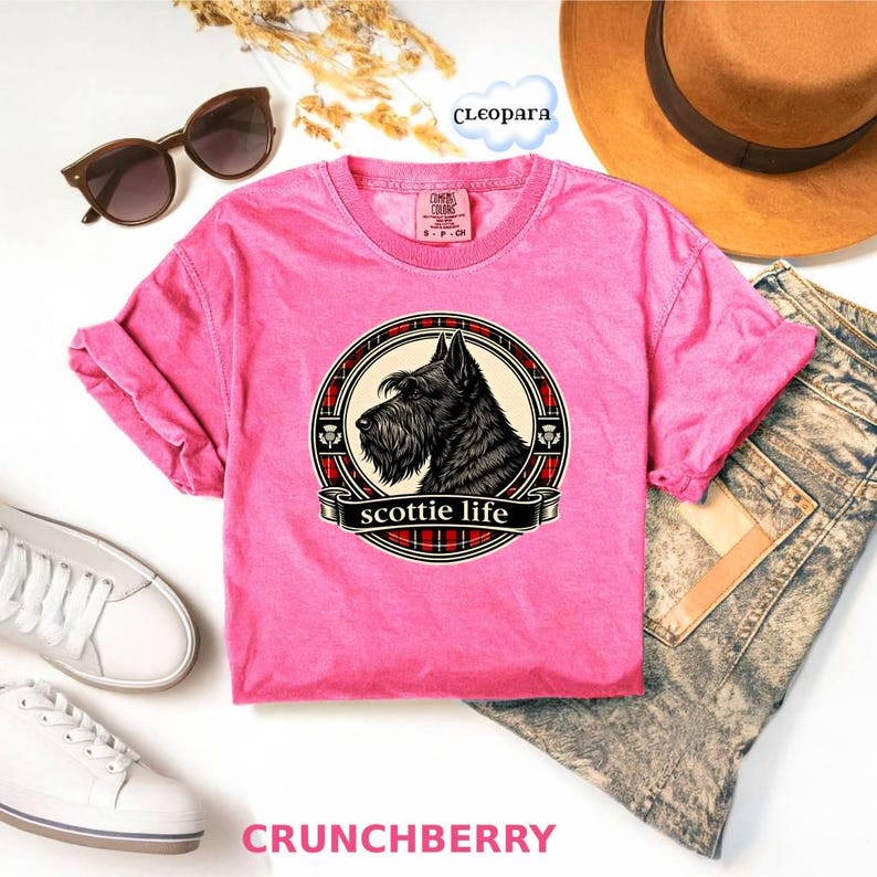 May include: A pink t-shirt with a graphic of a black Scottish Terrier dog in a circular design. The design includes the words "scottie life" on a banner. The shirt is paired with sunglasses, a hat, and jeans.