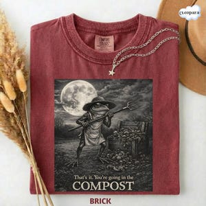 May include: Brick-colored t-shirt featuring a whimsical illustration of a frog in a hat and apron holding a pitchfork, with the text "That's it. You're going in the COMPOST." The shirt has a crew neck and short sleeves.