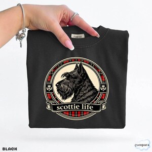 May include: Black crew neck sweatshirt featuring a circular graphic of a Scottish Terrier dog with the text "scottie life" on a banner. The graphic is framed by a red and black plaid pattern. The word "BLACK" is at the bottom left.