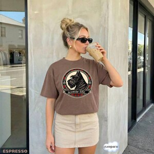 May include: Brown t-shirt with a circular graphic featuring a black Scottish Terrier dog and the text "scottie life". The graphic is framed with a red and black plaid pattern. The t-shirt is worn by a person.