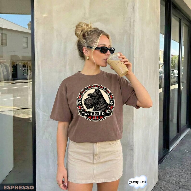 May include: Brown t-shirt with a circular graphic featuring a black Scottish Terrier dog and the text "scottie life". The graphic is framed with a red and black plaid pattern. The t-shirt is worn by a person.