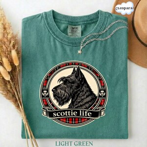 May include: Light green t-shirt with a graphic of a black Scottish Terrier dog in a circular design with red plaid accents and the words "scottie life". A silver necklace with a star pendant is draped on the shirt.