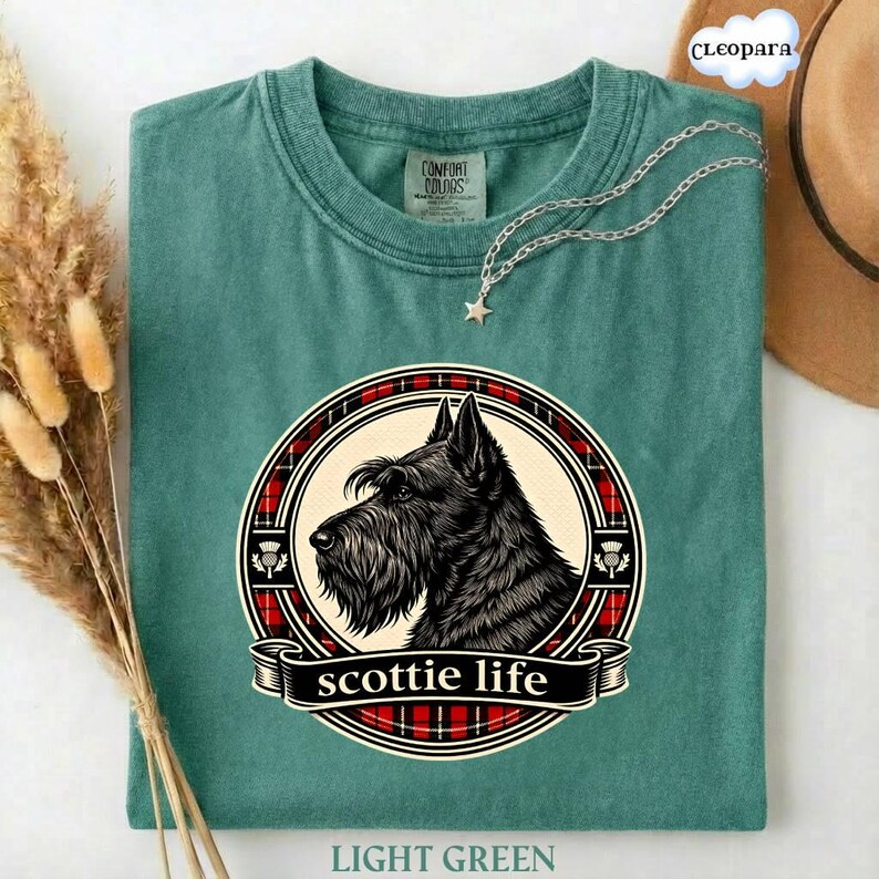May include: Light green t-shirt with a graphic of a black Scottish Terrier dog in a circular design with red plaid accents and the words "scottie life". A silver necklace with a star pendant is draped on the shirt.