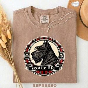May include: Espresso-colored t-shirt featuring a graphic of a Scottish Terrier dog within a circular design with a red and black plaid border. The text "scottie life" is displayed on a banner below the dog.