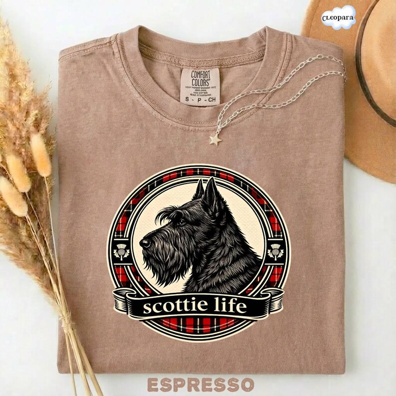 May include: Espresso-colored t-shirt featuring a graphic of a Scottish Terrier dog within a circular design with a red and black plaid border. The text "scottie life" is displayed on a banner below the dog.