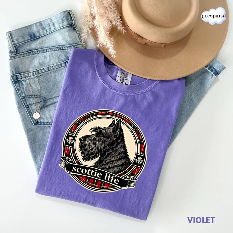 May include: A violet t-shirt featuring a black Scottie dog illustration within a circular design with a red and black plaid border. The words "scottie life" are on a banner below the dog. The shirt is paired with light blue jeans and a tan hat.