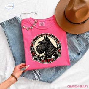 May include: A pink t-shirt featuring a black Scottish Terrier illustration within a circular design with a plaid border and the words "scottie life". The shirt is paired with blue jeans and a brown hat.