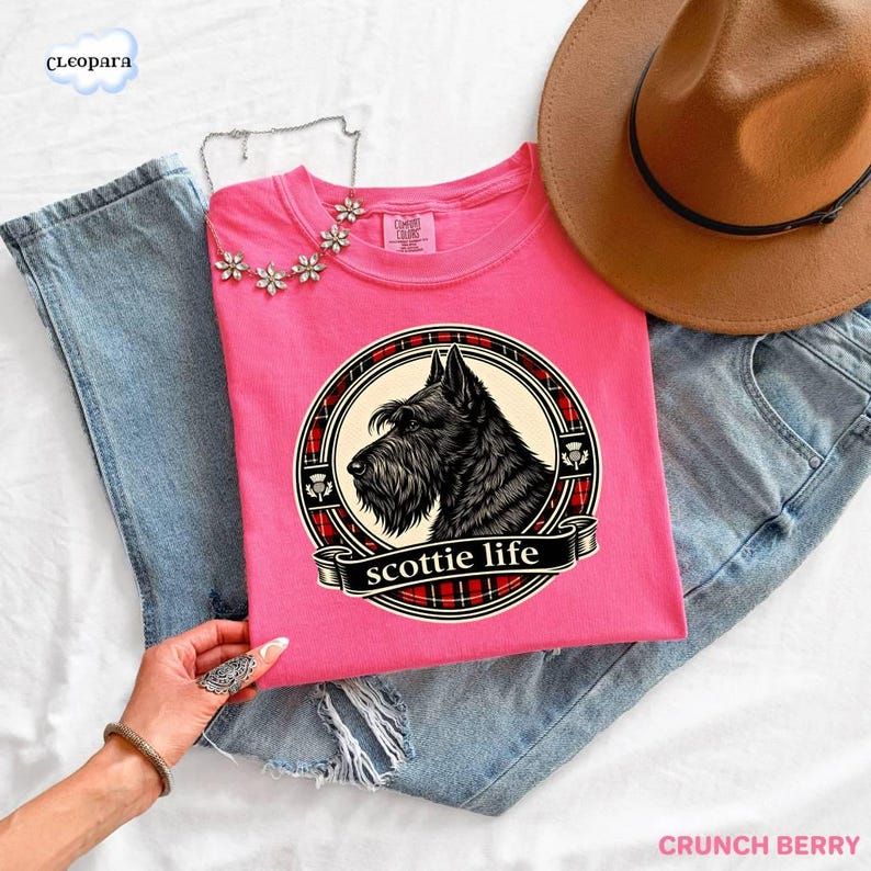 May include: A pink t-shirt featuring a black Scottish Terrier illustration within a circular design with a plaid border and the words "scottie life". The shirt is paired with blue jeans and a brown hat.