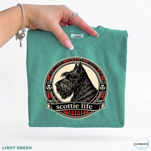 May include: Light green t-shirt featuring a black Scottie dog illustration within a red and black plaid circle. The words "scottie life" are displayed on a banner below the dog. The shirt is a solid light green color.