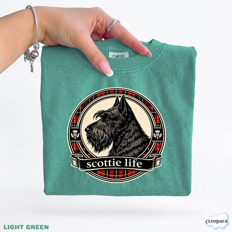 May include: Light green t-shirt featuring a black Scottie dog illustration within a red and black plaid circle. The words "scottie life" are displayed on a banner below the dog. The shirt is a solid light green color.