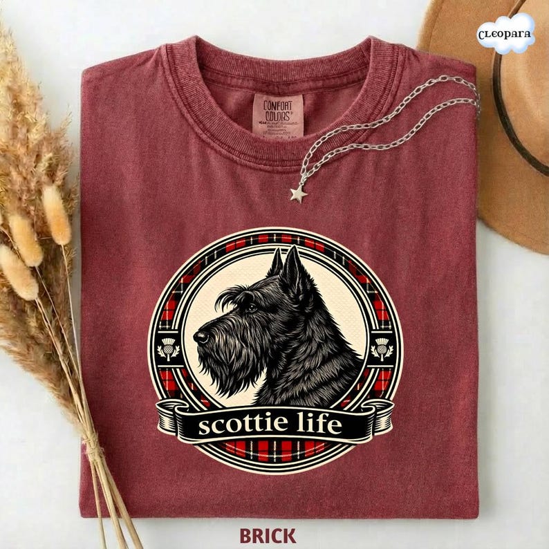 May include: Brick-colored t-shirt featuring a circular graphic of a Scottish Terrier dog with the words "scottie life" on a banner. The graphic is surrounded by a red and black plaid pattern. A silver necklace with a star pendant is draped on the shirt.