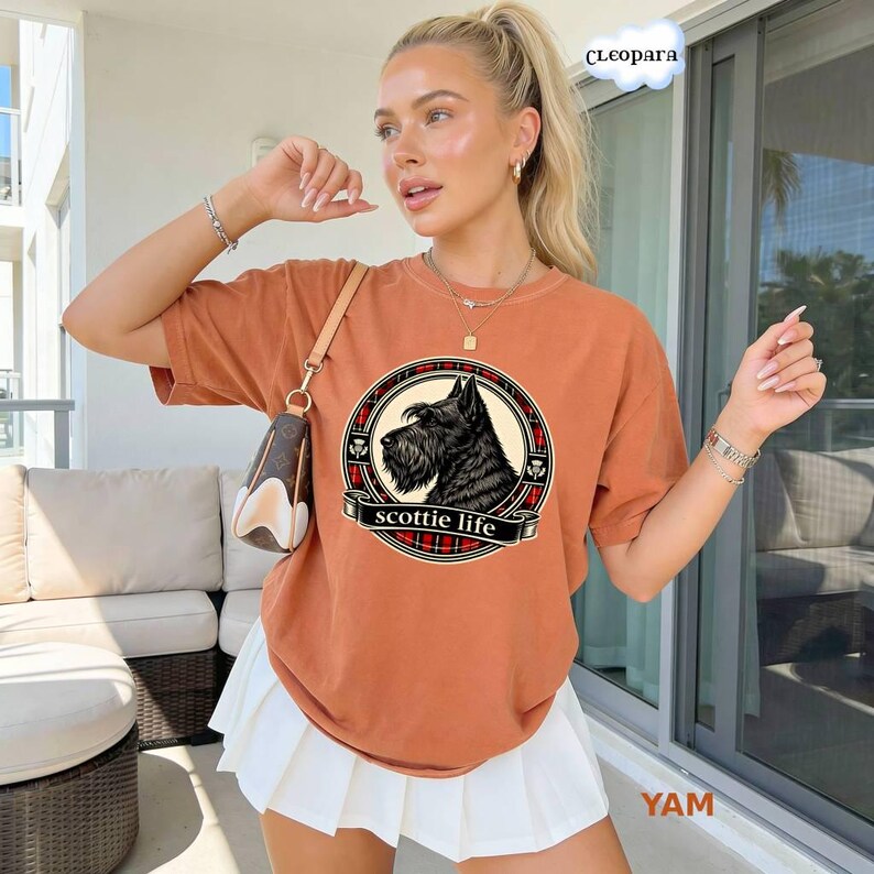 May include: Burnt orange t-shirt featuring a black and white illustration of a Scottish Terrier dog within a circular design. The design includes a red and black plaid pattern and the words "scottie life".