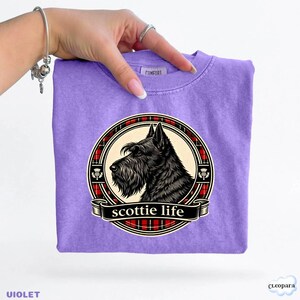 May include: A violet t-shirt featuring a black Scottie dog illustration within a circular design. The design includes a red and black plaid border and the words "scottie life" on a banner. The shirt is held by a hand.