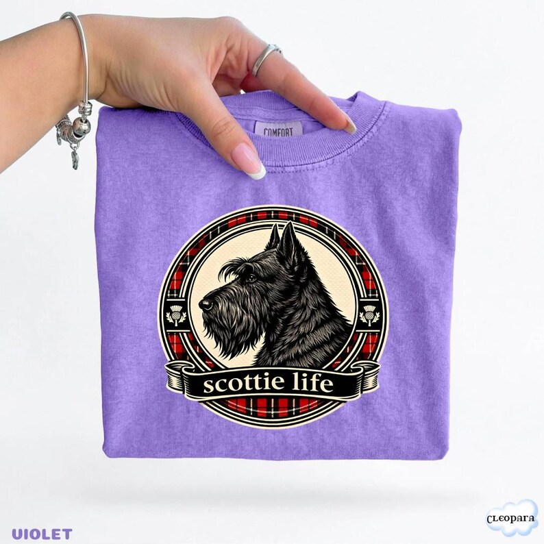 May include: A violet t-shirt featuring a black Scottie dog illustration within a circular design. The design includes a red and black plaid border and the words "scottie life" on a banner. The shirt is held by a hand.