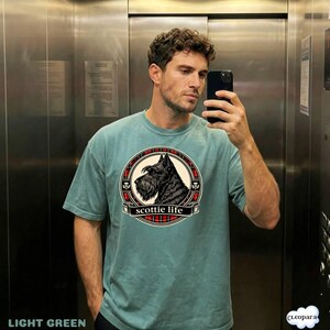 May include: Light green t-shirt featuring a circular graphic of a Scottish Terrier dog with the text "scottie life". The graphic is framed by a red and black plaid pattern. The shirt is a solid light green color.